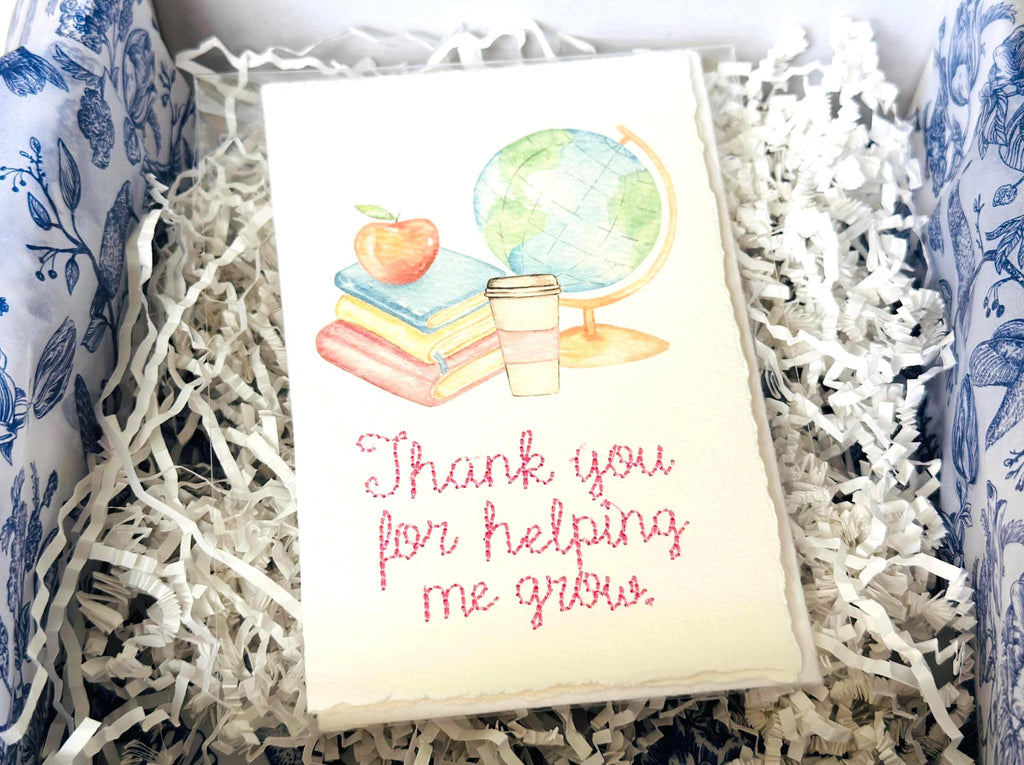 A handmade card that has watercolor art of a pile of books with an apple on top next to a globe and a to-go cup of coffee in soft pastel colors with embroidery under it that says: Thank you for helping me grow. The embroidery is done in pink thread. The card is in a box with shredded paper and chinoiserie tissue.