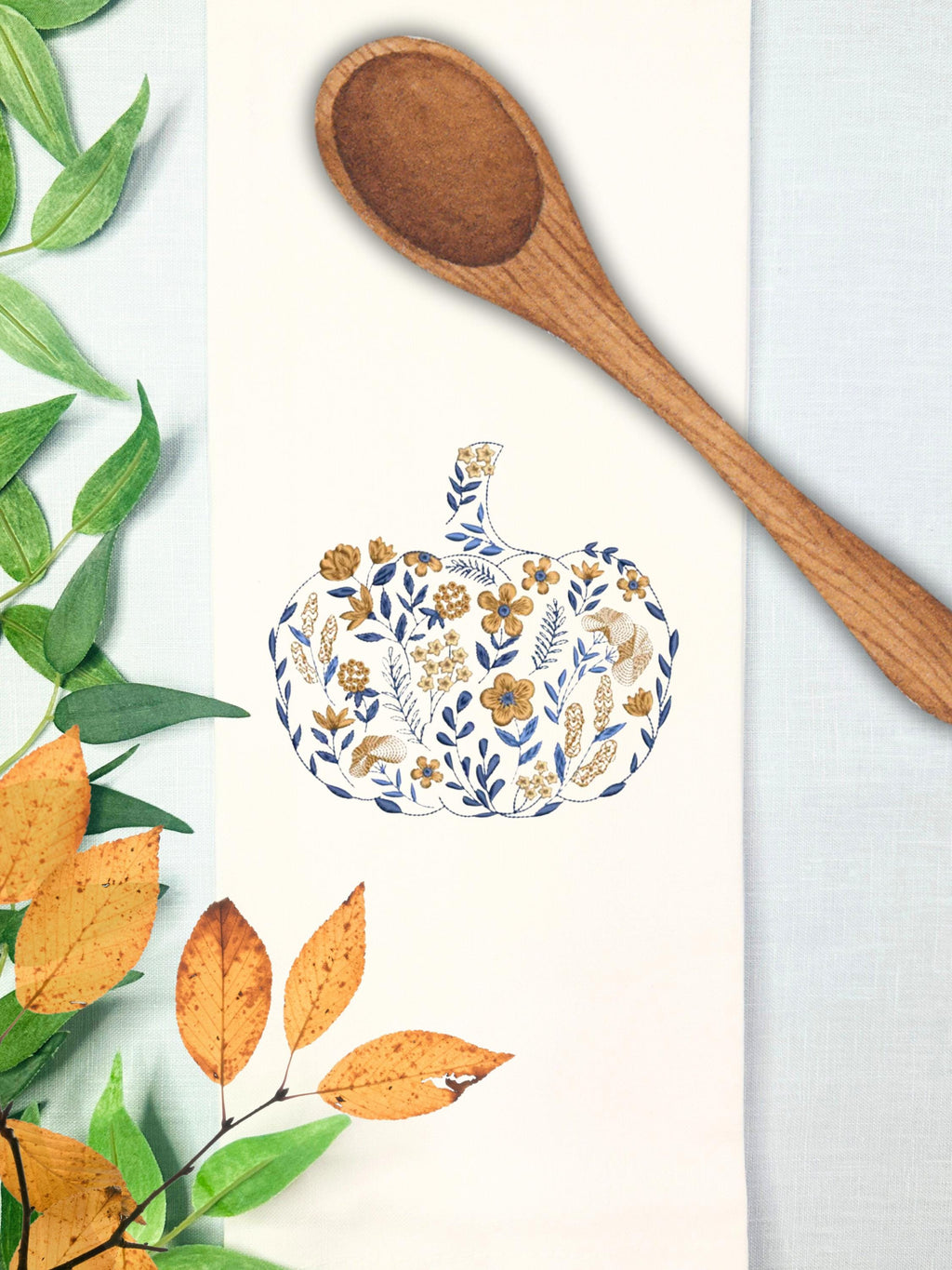 A blue and orange pumpkin that is embroidered in a chinoiserie coquette style filled with flowers of all different sizes on a white tea towel measuring 17x30&quot; is on a light blue linen tablecloth with green and orange leaves to the left and a wooden spoon on the top.