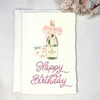 A deckled edge card with watercolor art of a champagne bottle with a pink bow and pink flowers to the left. Under the art is embroidered: Happy Birthday in pink. The card is set on a white linen table with pink cherry blossoms to the top right. Cards are approximately 4x6.
