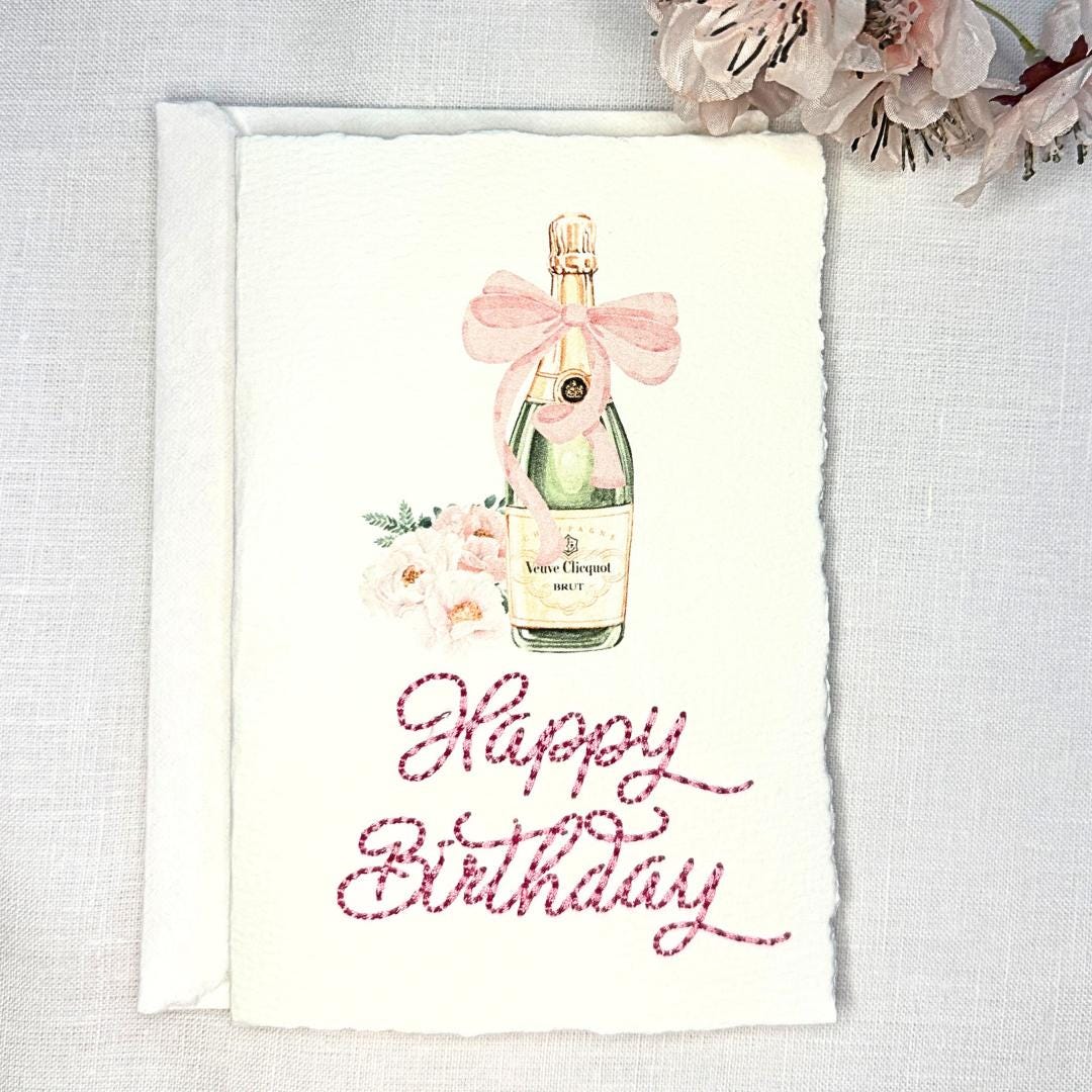 A deckled edge card with watercolor art of a champagne bottle with a pink bow and pink flowers to the left. Under the art is embroidered: Happy Birthday in pink. The card is set on a white linen table with pink cherry blossoms to the top right. Cards are approximately 4x6.