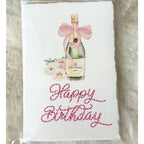 A deckled edge card with watercolor art of a champagne bottle with a pink bow and pink flowers to the left. Under the art is embroidered: Happy Birthday in pink. The card is set on a white fluffy blanket. Cards are approximately 4x6.