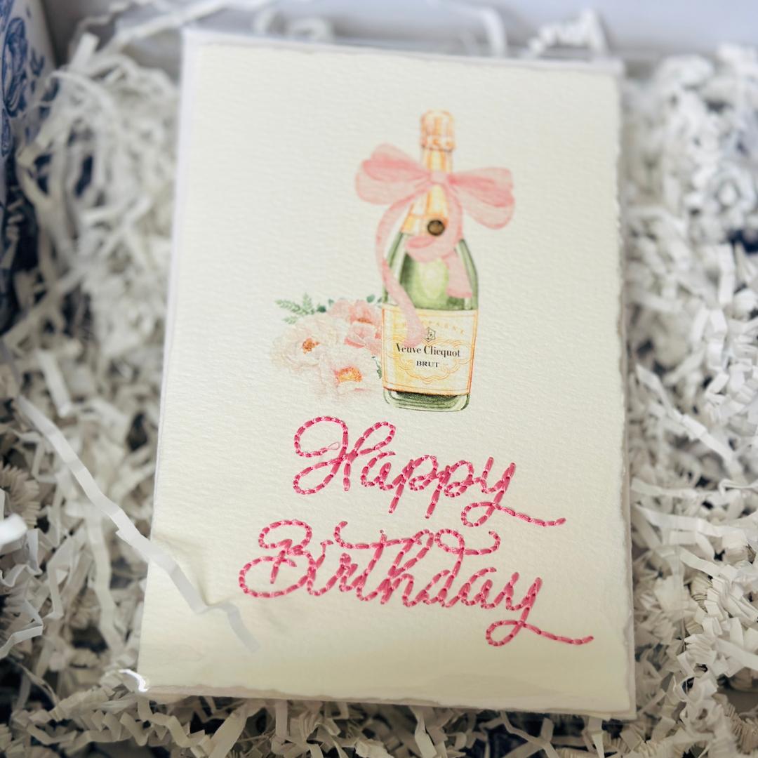 A deckled edge card with watercolor art of a champagne bottle with a pink bow and pink flowers to the left. Under the art is embroidered: Happy Birthday in pink. The card is set on white crinkle paper with blue and white chinoiserie tissue. Cards are approximately 4x6.