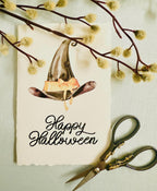A photo of a handmade card that has a watercolor witch&#39;s hat in black with an orange bow. Under the hat the card is embroidered in black thread &quot;Happy Halloween&quot;. The card is set on a light blue linen tablecloth with a pair of embroidery scissors and a branch with white, puffy buds.