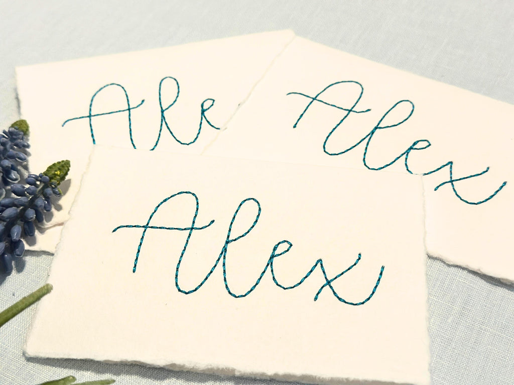 Three handmade cards with the name Alex embroidered onto them in teal. The cards are set on a blue linen tablecloth and purple cone flowers.