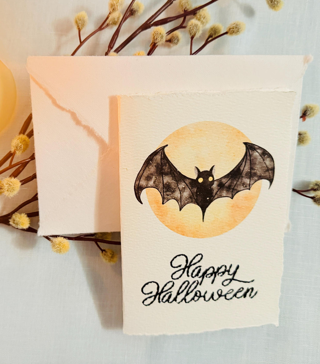 A watercolor bat in front of a moon with Happy Halloween embroidered below. The card is sitting on top of its&#39; matching envelope which is laying on top of a branch with fuzzy white buds on a blue linen tablecloth. Cards are approximately 4&quot;x6&quot;.