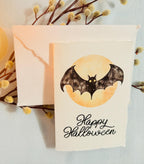 A watercolor bat in front of a moon with Happy Halloween embroidered below. The card is sitting on top of its&#39; matching envelope which is laying on top of a branch with fuzzy white buds on a blue linen tablecloth. Cards are approximately 4&quot;x6&quot;.