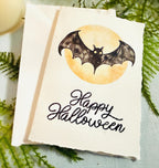 A watercolor bat in front of a moon with Happy Halloween embroidered below. The card is sitting on top of its&#39; matching envelope which is laying on top of a fern on a blue linen tablecloth. Cards are approximately 4&quot;x6&quot;. There is a candle to the upper left.