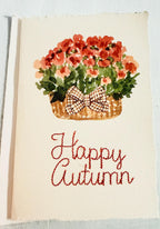 Watercolor orange mums with greenery and a plaid brown bow in a mid-tone wicker basket with &quot;Happy Autumn&quot; embroidered below in orange. The card is set on top of its&#39; envelope on a blue linen tablecloth.
