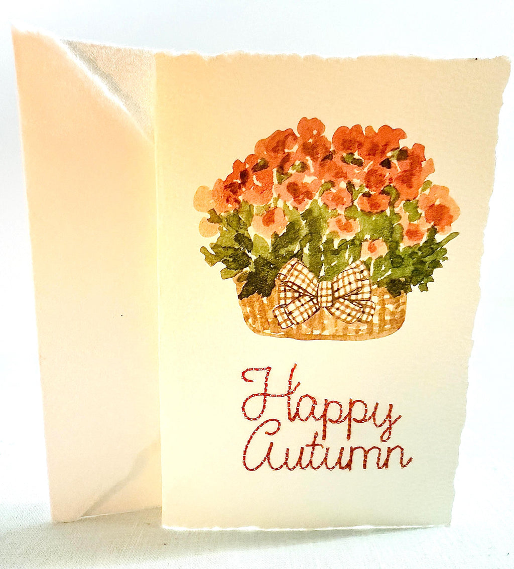 Watercolor orange mums with greenery and a plaid brown bow in a mid-tone wicker basket with &quot;Happy Autumn&quot; embroidered below in orange.The card is set on top of its&#39; envelope on a blue linen tablecloth.