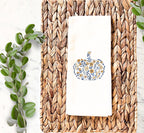 Beautiful blue and golden orange Embroidered Pumpkin on a white tea towel, on top of a marble countertop with a wicker placemat  and pretty greens to the left used for Fall Hosting Decor, Thanksgiving Towel, Hostess Gifts, for Your Best Friend.