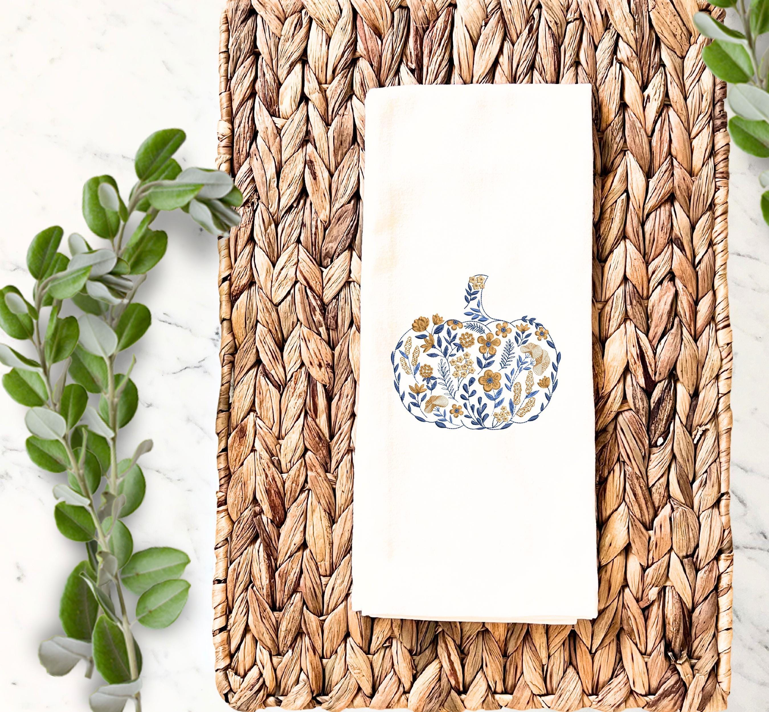 Beautiful blue and golden orange Embroidered Pumpkin on a white tea towel, on top of a marble countertop with a wicker placemat  and pretty greens to the left used for Fall Hosting Decor, Thanksgiving Towel, Hostess Gifts, for Your Best Friend.