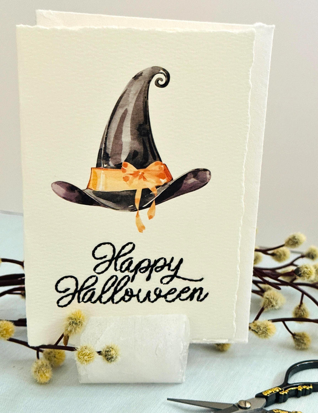 A photo of a handmade card that has a watercolor witch&#39;s hat in black with an orange bow. Under the hat the card is embroidered in black thread &quot;Happy Halloween&quot;. The card is set in a stone card holder on top of a light blue linen tablecloth with a wooden branch with white puffy buds.