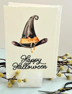A photo of a handmade card that has a watercolor witch&#39;s hat in black with an orange bow. Under the hat the card is embroidered in black thread &quot;Happy Halloween&quot;. The card is set in a stone card holder on top of a light blue linen tablecloth with a wooden branch with white puffy buds.