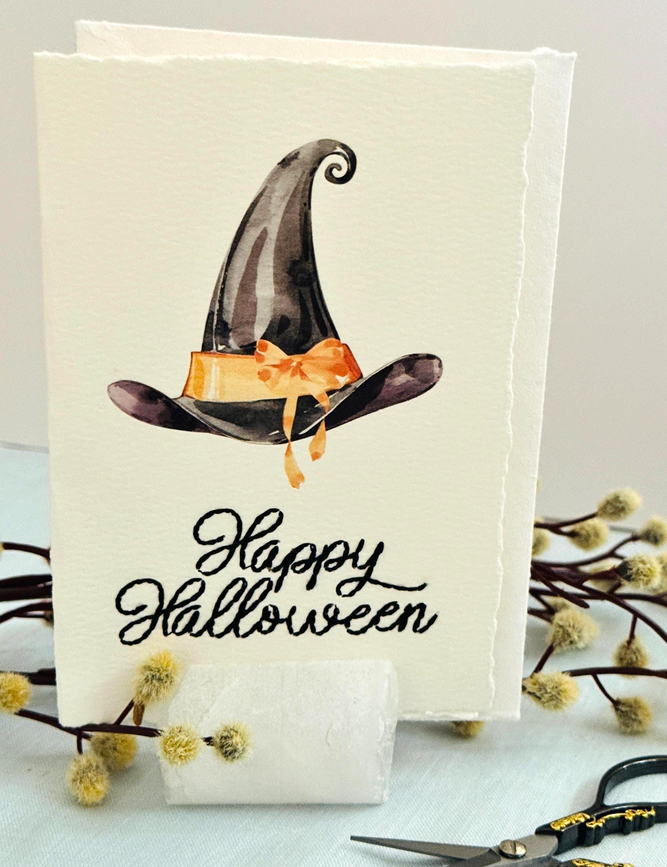 A photo of a handmade card that has a watercolor witch&#39;s hat in black with an orange bow. Under the hat the card is embroidered in black thread &quot;Happy Halloween&quot;. The card is set in a stone card holder on top of a light blue linen tablecloth with a wooden branch with white puffy buds.