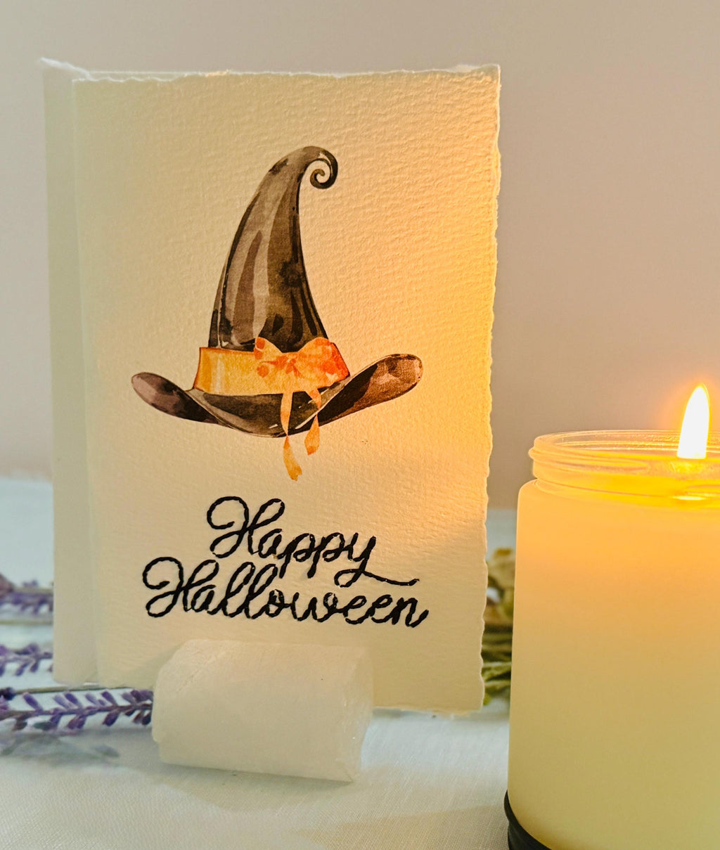 A photo of a handmade card that has a watercolor witch&#39;s hat in black with an orange bow. Under the hat the card is embroidered in black thread &quot;Happy Halloween&quot;. The card is set in a stone card holder on top of a light blue linen tablecloth with a lit candle next to it and a purple lavender sprig behind it.