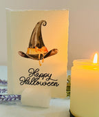 A photo of a handmade card that has a watercolor witch&#39;s hat in black with an orange bow. Under the hat the card is embroidered in black thread &quot;Happy Halloween&quot;. The card is set in a stone card holder on top of a light blue linen tablecloth with a lit candle next to it and a purple lavender sprig behind it.