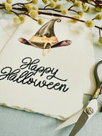 A photo of a handmade card that has a watercolor witch&#39;s hat in black with an orange bow. Under the hat the card is embroidered in black thread &quot;Happy Halloween&quot;. The card is set on top of a light blue linen tablecloth with embroidery scissors to the right and a sprig of wood with puffy white buds.
