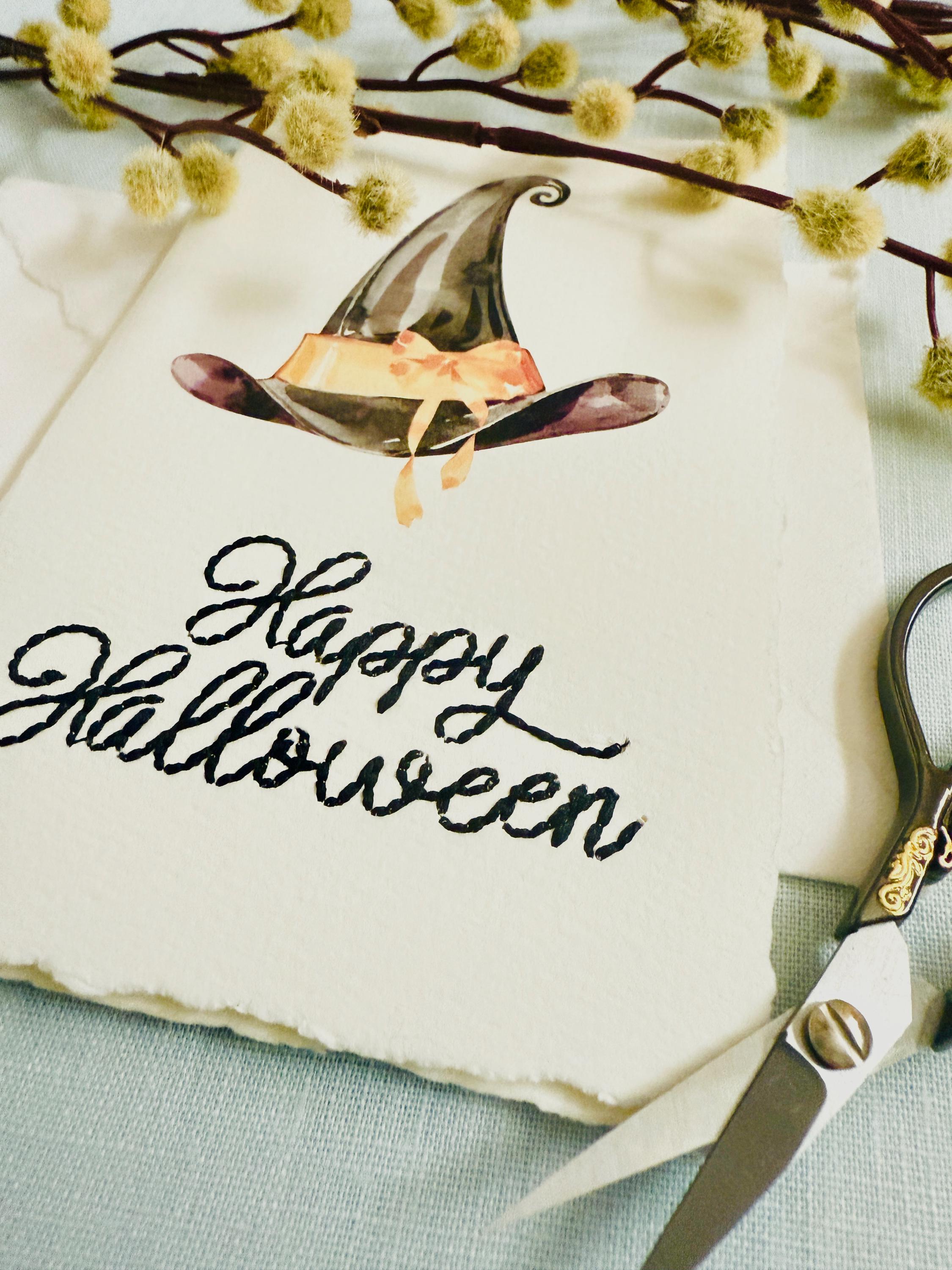 A photo of a handmade card that has a watercolor witch&#39;s hat in black with an orange bow. Under the hat the card is embroidered in black thread &quot;Happy Halloween&quot;. The card is set on top of a light blue linen tablecloth with embroidery scissors to the right and a sprig of wood with puffy white buds.