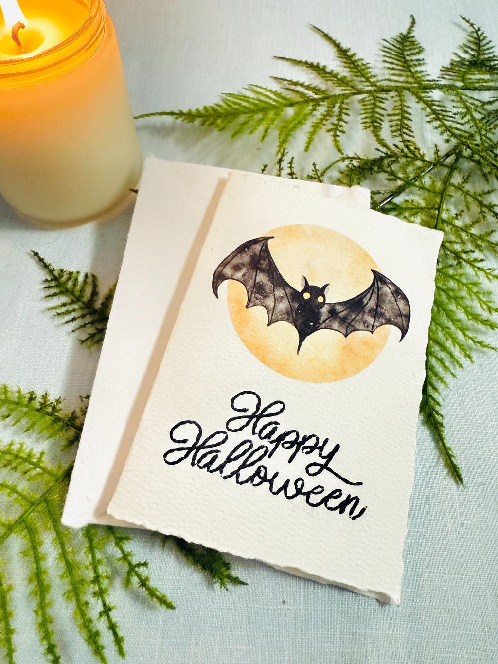 A watercolor bat in front of a moon with Happy Halloween embroidered below. The card is sitting on top of its&#39; matching envelope which is laying on top of a fern on a blue linen tablecloth. Cards are approximately 4&quot;x6&quot;. There is a candle to the upper right.
