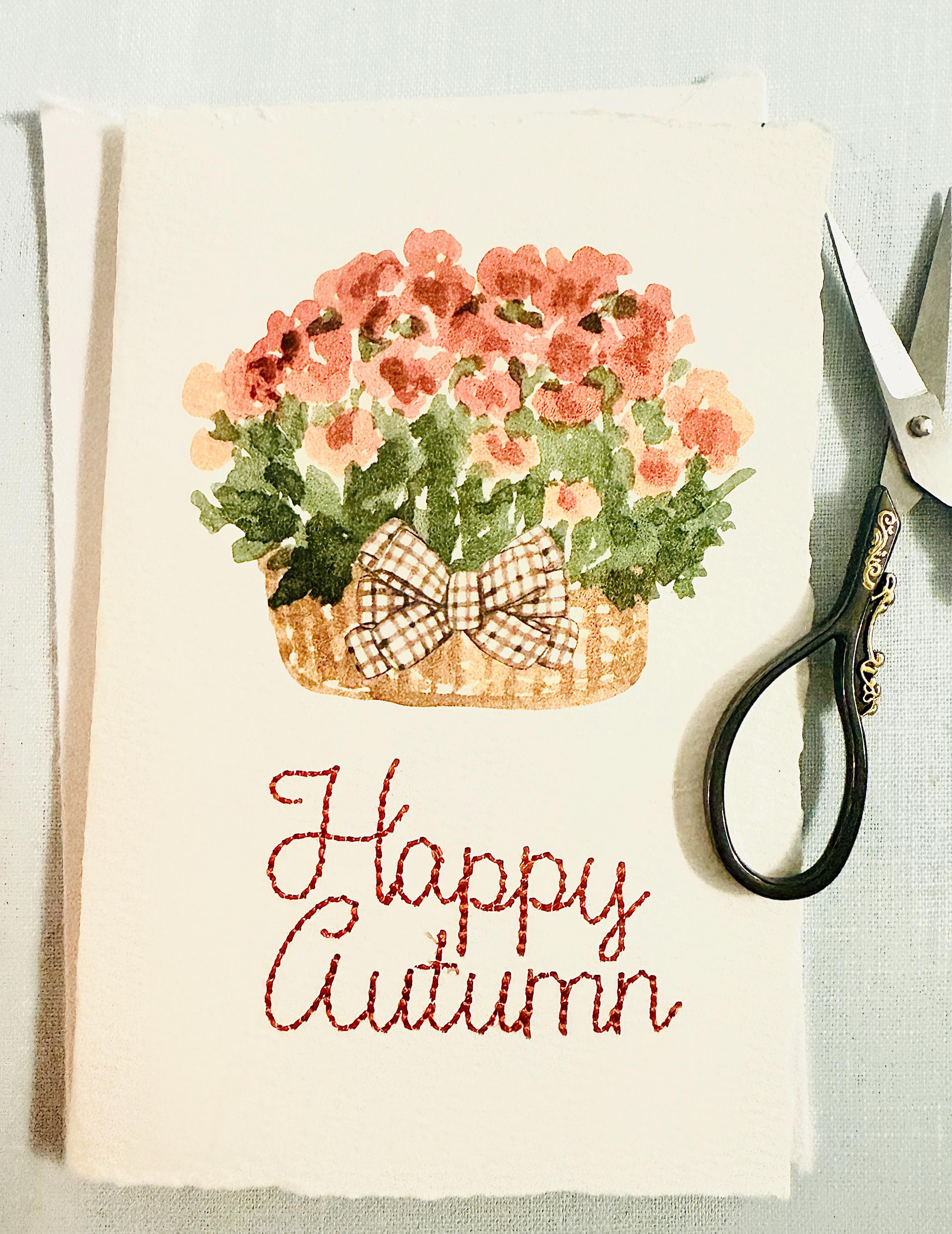 Watercolor orange mums with greenery and a plaid brown bow in a mid-tone wicker basket with &quot;Happy Autumn&quot; embroidered below in orange. There are embroidery scissors to the right that have black handles with gold embellishment. The card is set on top of its&#39; envelope on a blue linen tablecloth.