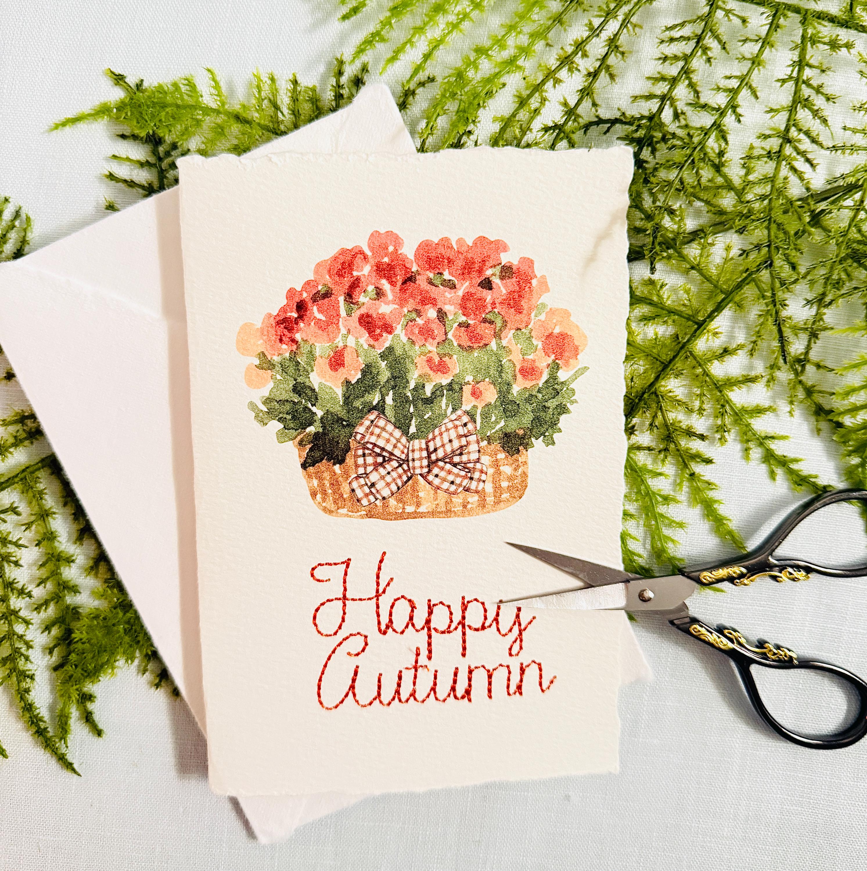 Watercolor orange mums with greenery and a plaid brown bow in a mid-tone wicker basket with &quot;Happy Autumn&quot; embroidered below in orange. There are embroidery scissors to the right that have black handles with gold embellishment. The card is set on top of its&#39; envelope on a blue linen tablecloth and a fern.
