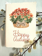 Watercolor orange mums with greenery and a plaid brown bow in a mid-tone wicker basket with &quot;Happy Autumn&quot; embroidered below in orange. The card is set on top of a branch with white, puffy bud on a light blue linen tablecloth.