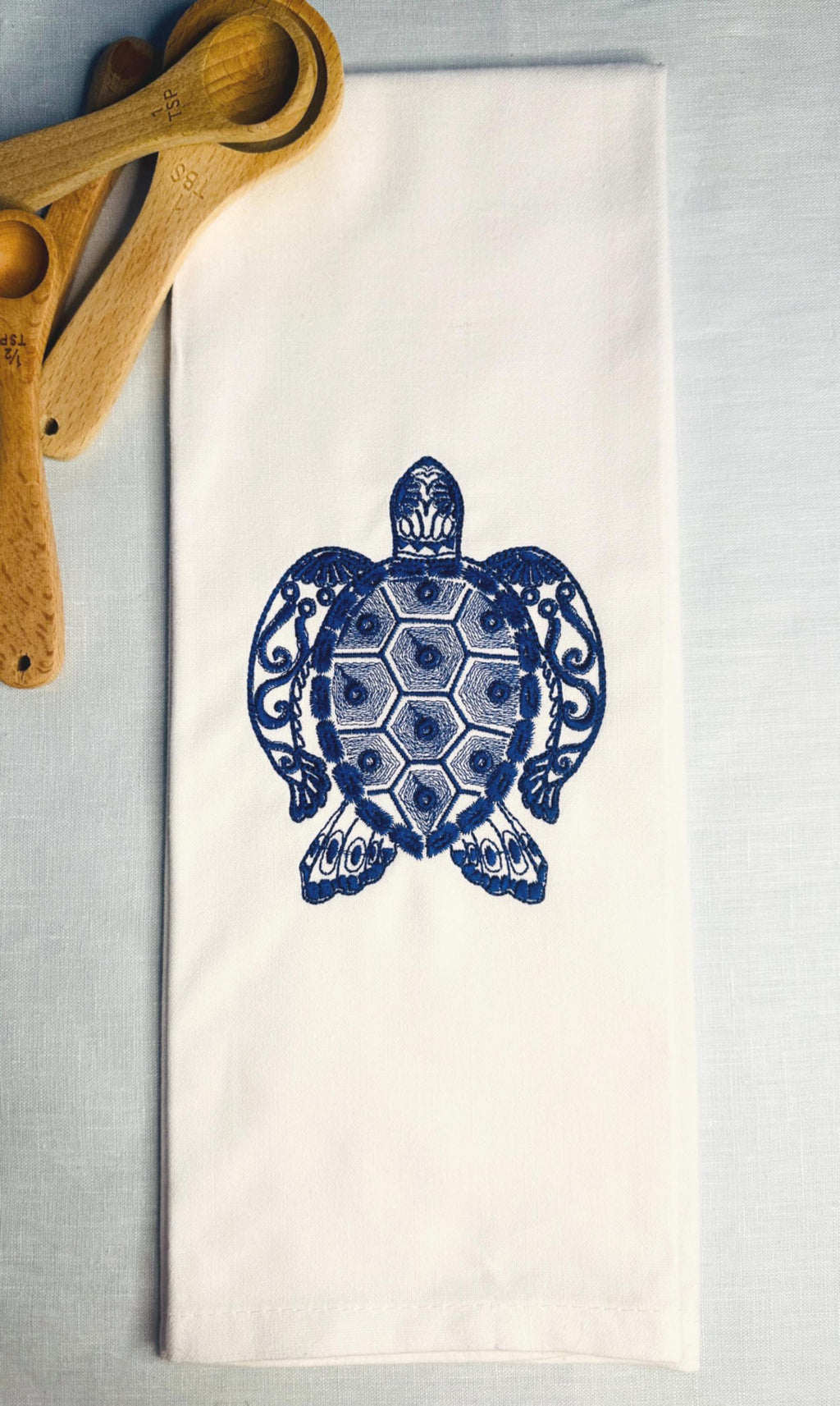 Coastal decor of a sea turtle embroidered in blue on a white tea towel set on a blue linen background with wooden measuring spoons in the upper left corner.