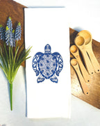 Sea Turtle Ocean Themed tea towel stitched in blue on a white tea towel set on a cutting board. There are flowers to the left and measuring spoons to the right.
