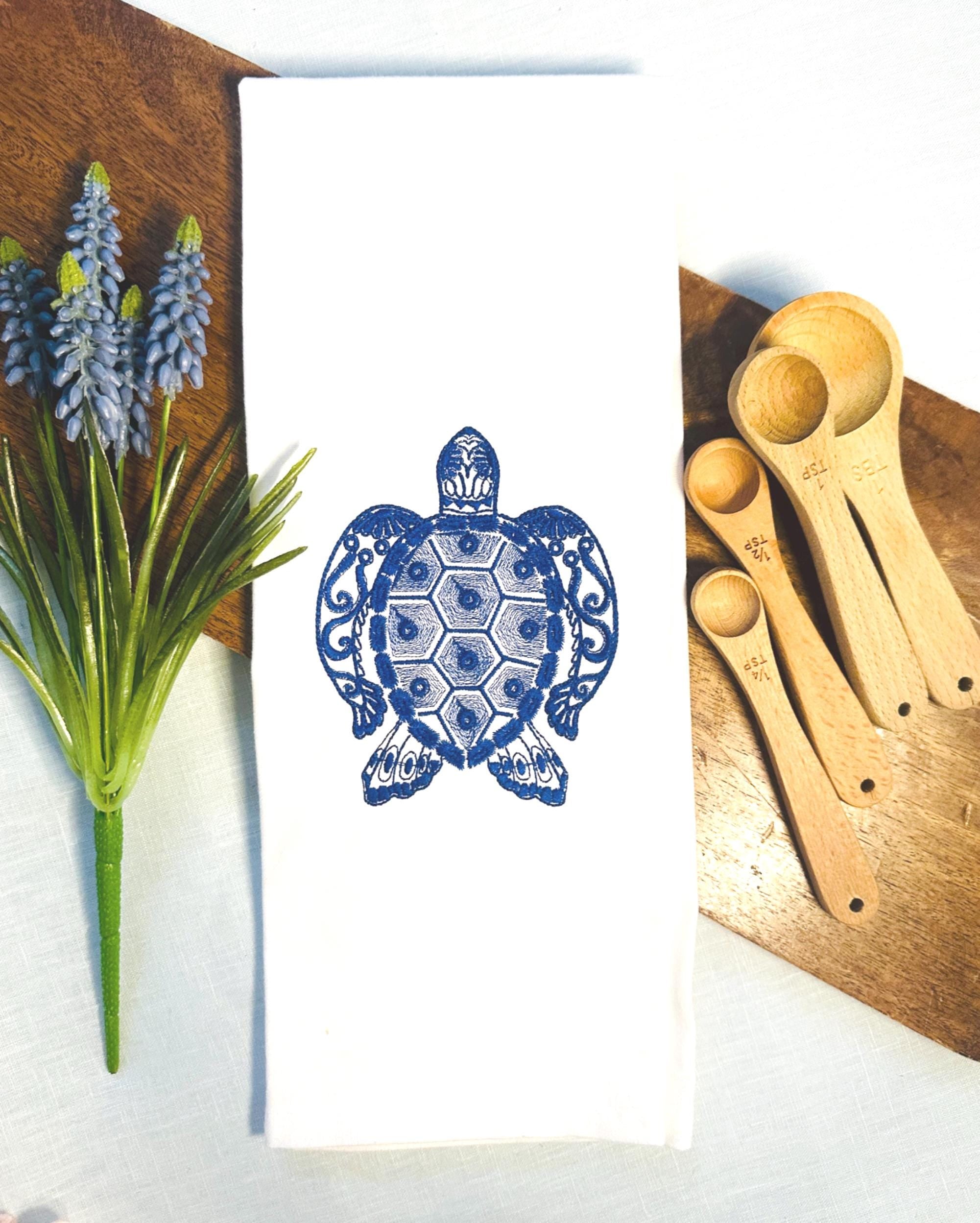 Sea Turtle Ocean Themed tea towel stitched in blue on a white tea towel set on a cutting board. There are flowers to the left and measuring spoons to the right.