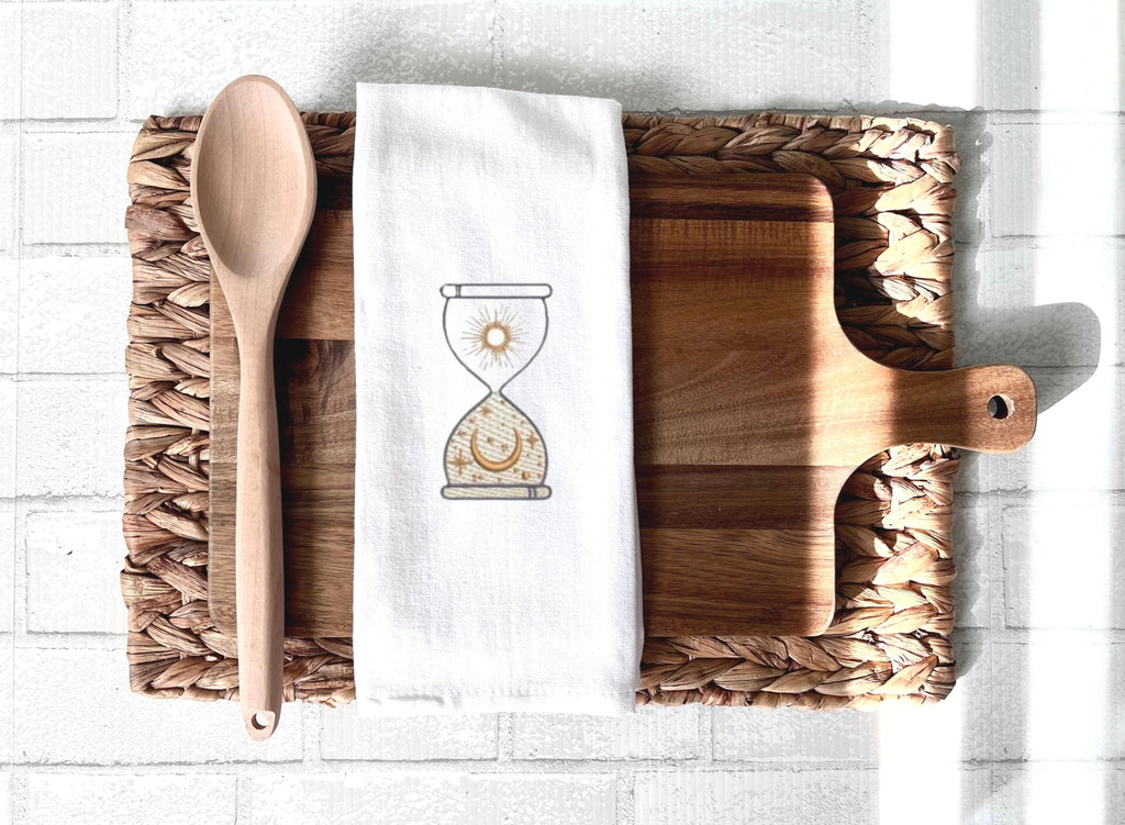 An image of an hourglass with sun and moon embroidered in gold on a white tea towel set on a cutting board with a spoon on top of a woven placemat on a white tile background.