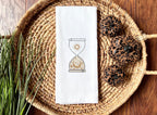 An image of an hourglass with sun and moon embroidered in gold on a white tea towel set in a basket with cheetah design pumpkins to the right and greens on the left set on a wooden table.