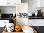 White tea towel on a towel holder with Happy Halloween embroidered in black. The word happy is inside the bat. It is set on a kitchen counter with a tray with black handles under along with skeleton pumpkins and oils.