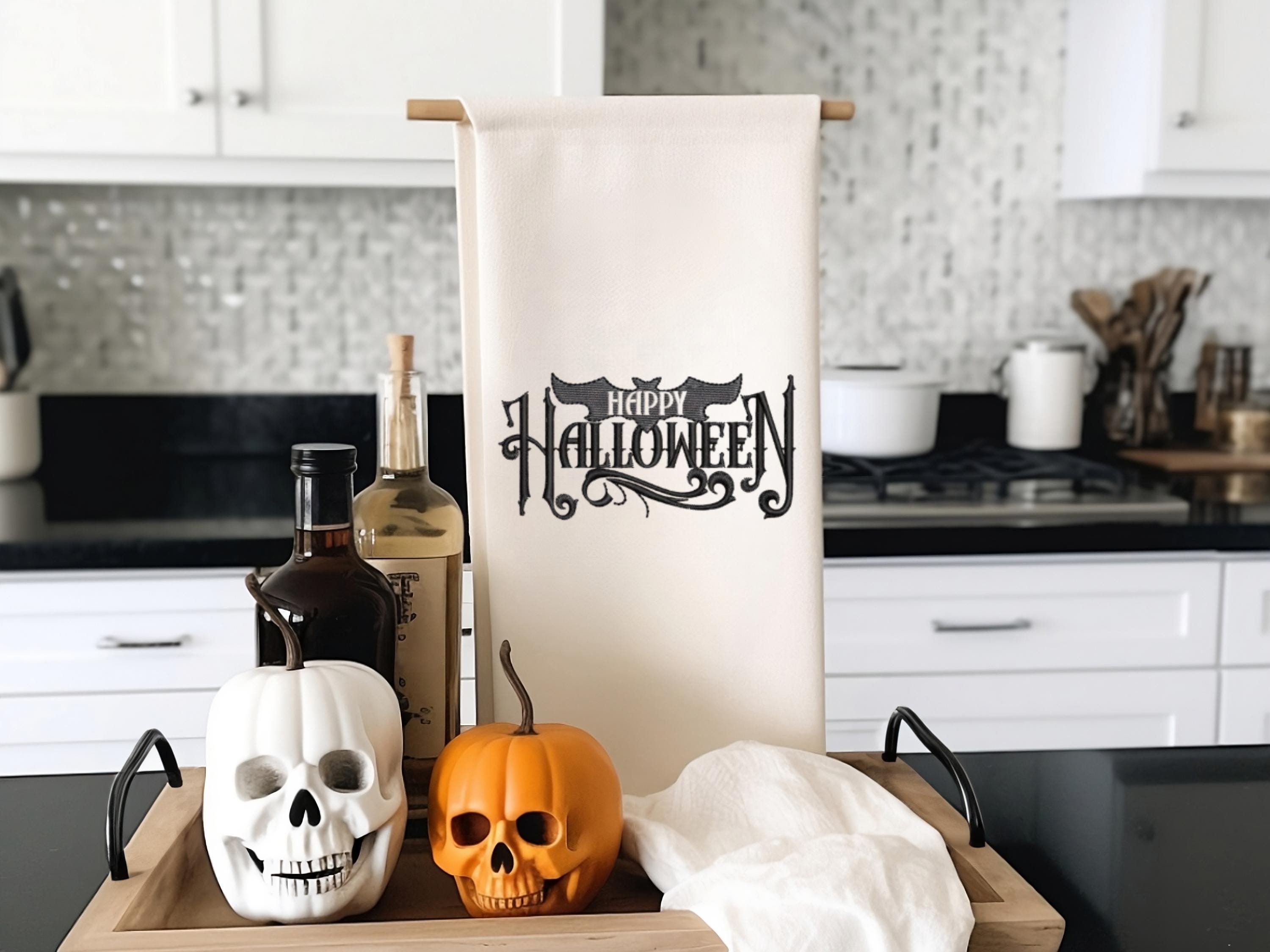 White tea towel on a towel holder with Happy Halloween embroidered in black. The word happy is inside the bat. It is set on a kitchen counter with a tray with black handles under along with skeleton pumpkins and oils.