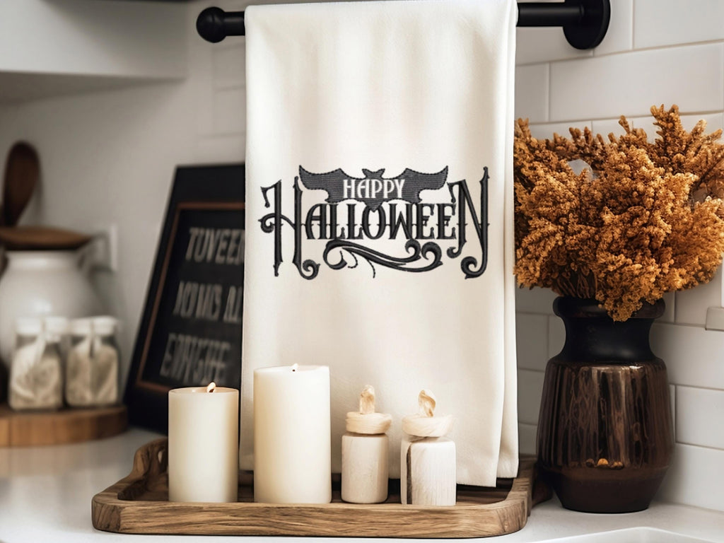 White tea towel on a black towel holder with Happy Halloween embroidered in black. The word happy is inside the bat. It is set on a kitchen counter with a tray under it and candles in front with mums to the right.