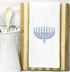 A menorah in different shades of blue embroidered on a white cotton tea towel set on a cutting board on a white marble kitchen counter with white subway backsplash and kitchen tools to the left in a white pitcher. Custom options are also available.