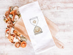 An image of an hourglass with sun and moon embroidered in gold on a white tea towel set on a wooden cutting board with dried oranges and spices to the left.