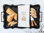 An image of an hourglass with sun and moon embroidered in gold on a white tea towel set on a black tray with bread and utensils and a few black branches to the bottom right.