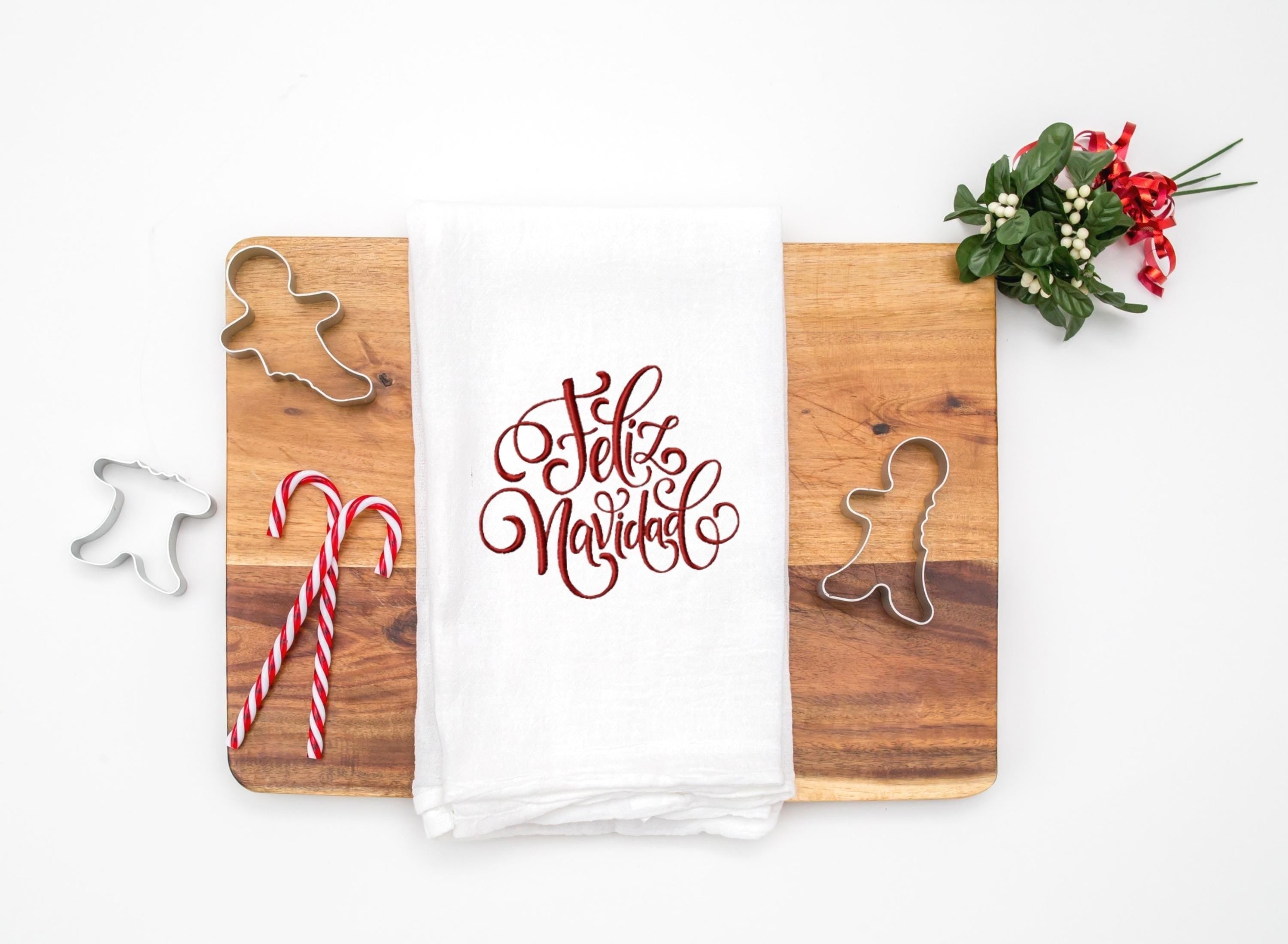 A white tea towel embroidered with Feliz Navidad in red on a cutting board with candy canes and cookie cutters and mistletoe to the top right. Set on a white table.