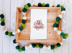 A white tea towel with Feliz Navidad embroidered in red on a wooden tray with a multi-color green and white garland on a white table.