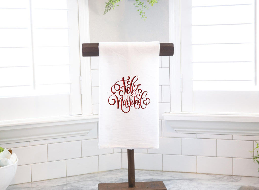 A white tea towel set on a towel holder on a white marble counter with white walls and windows behind. Feliz Navidad is embroidered in red on the towel.