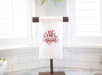 A white tea towel set on a towel holder on a white marble counter with white walls and windows behind. Feliz Navidad is embroidered in red on the towel.