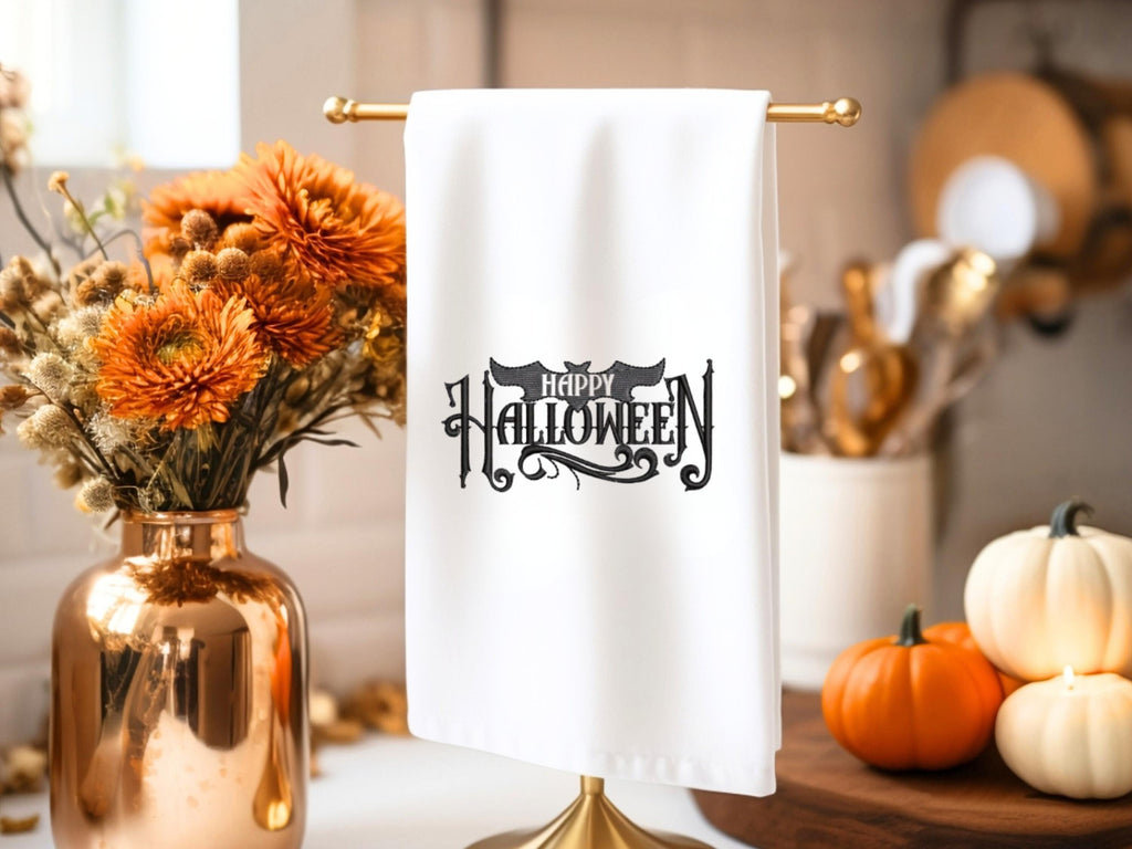 White tea towel on a copper towel holder with Happy Halloween embroidered in black. The word happy is inside the bat. It is set on a kitchen counter with a board behind it and mums to the left. There are little pumpkins on the board.