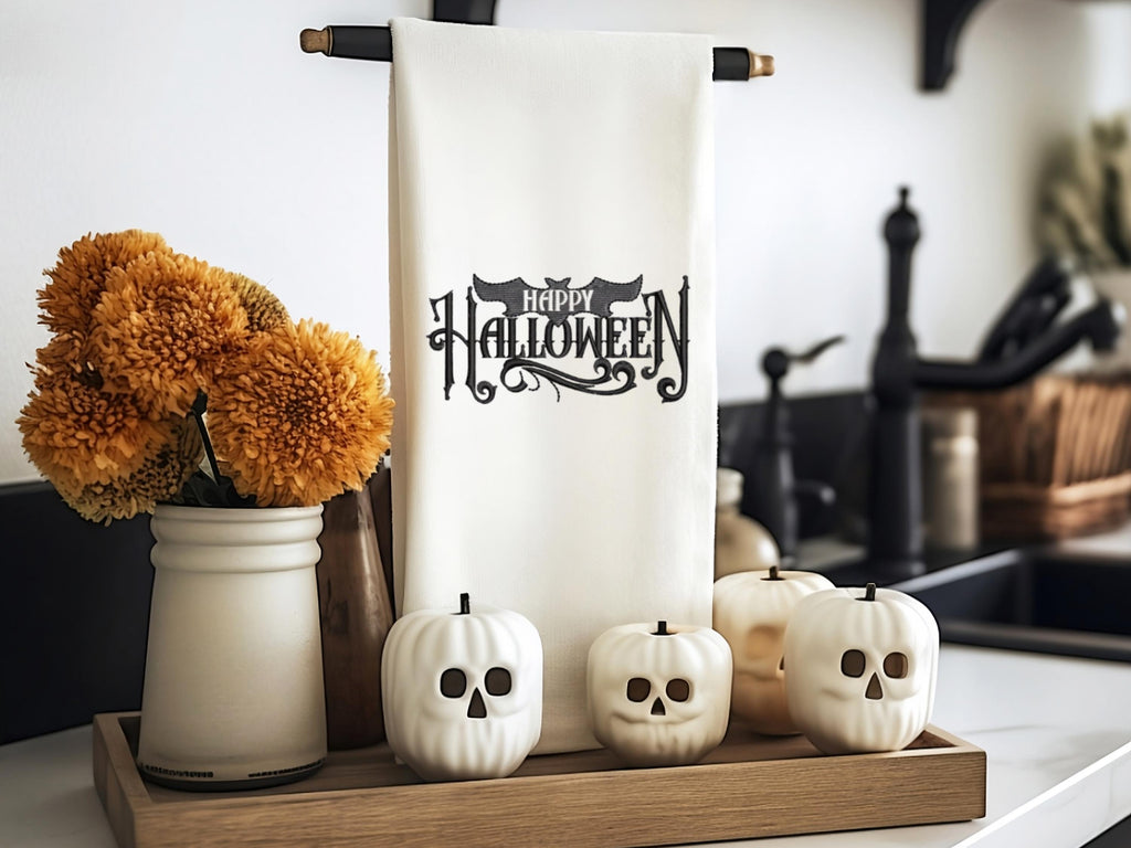 White tea towel on a black towel holder with Happy Halloween embroidered in black. The word happy is inside the bat. It is set on a kitchen counter with a board under it and mums to the left. There are little white pumpkins in front with skeleton faces.