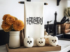 White tea towel on a black towel holder with Happy Halloween embroidered in black. The word happy is inside the bat. It is set on a kitchen counter with a board under it and mums to the left. There are little white pumpkins in front with skeleton faces.