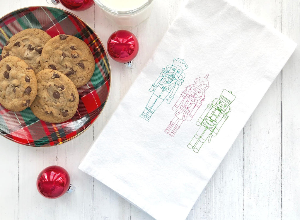 Three embroidered nutcracker figures on a white kitchen towel. The nutcrackers are outlined in green, pink, and blue thread. The nutcrackers are standing in a row and are facing the viewer.