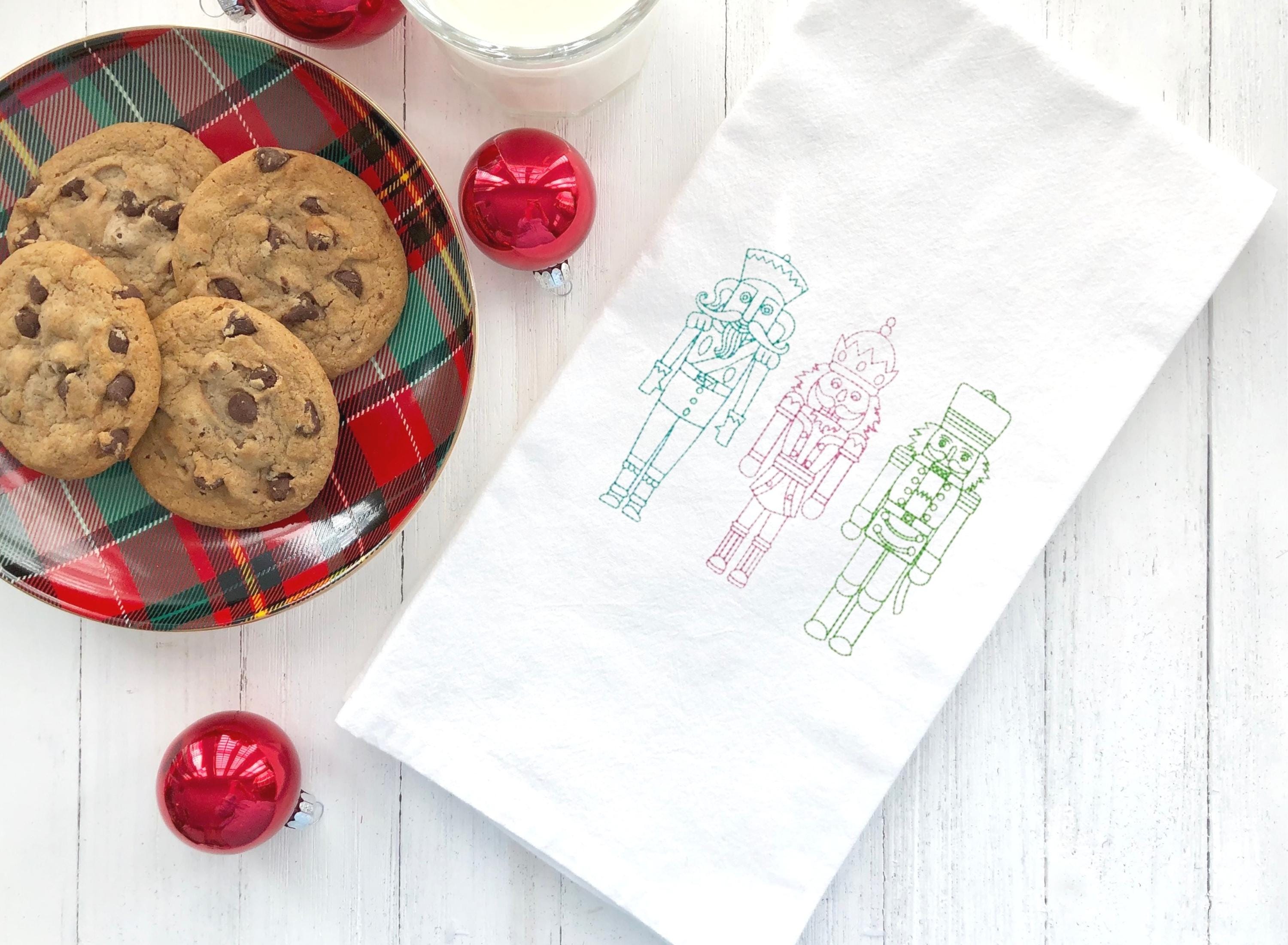 Three embroidered nutcracker figures on a white kitchen towel. The nutcrackers are outlined in green, pink, and blue thread. The nutcrackers are standing in a row and are facing the viewer.
