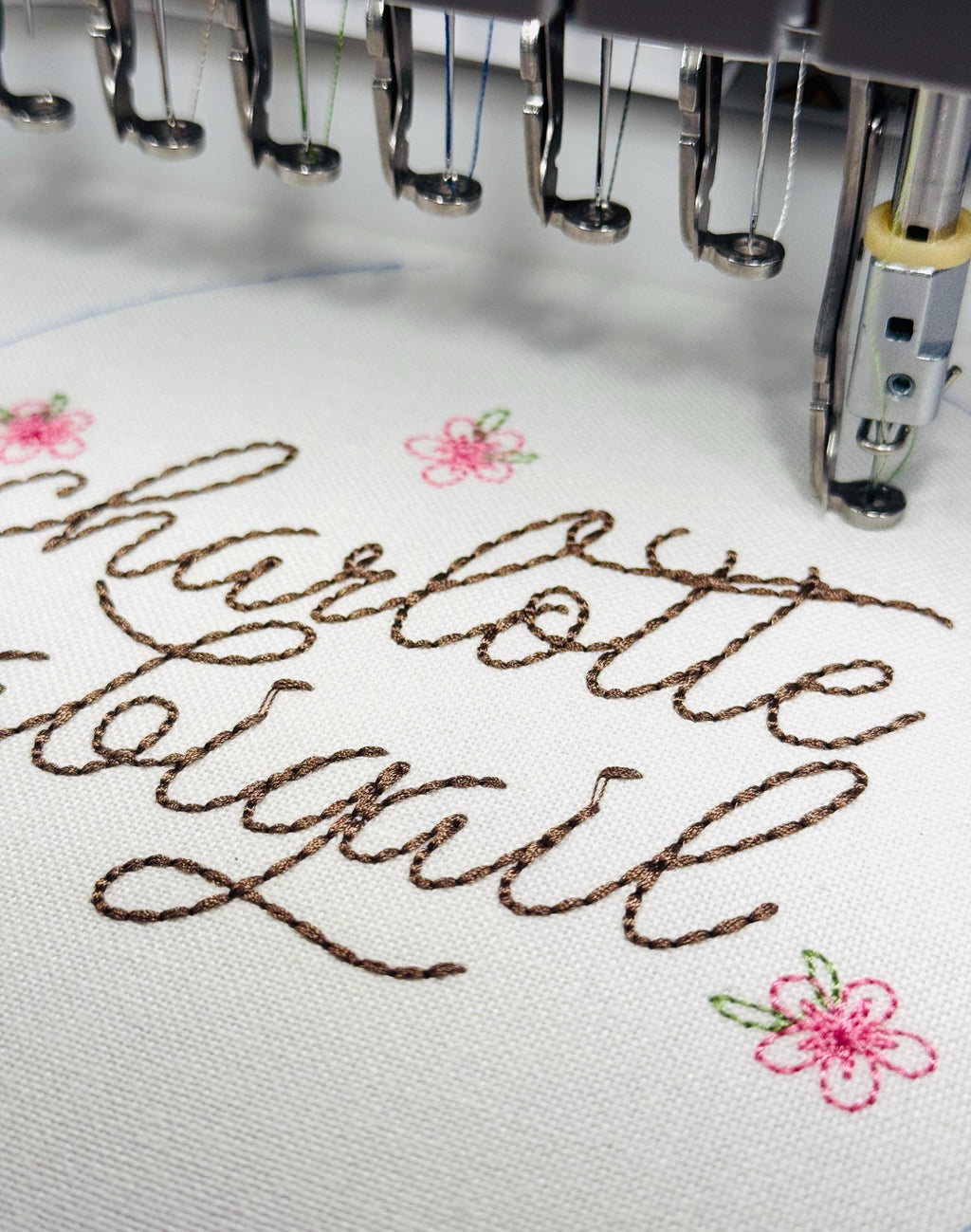 A close up of Charlotte Abigail in cursive being embroidered on white fabric. You can see three small pink flowers.