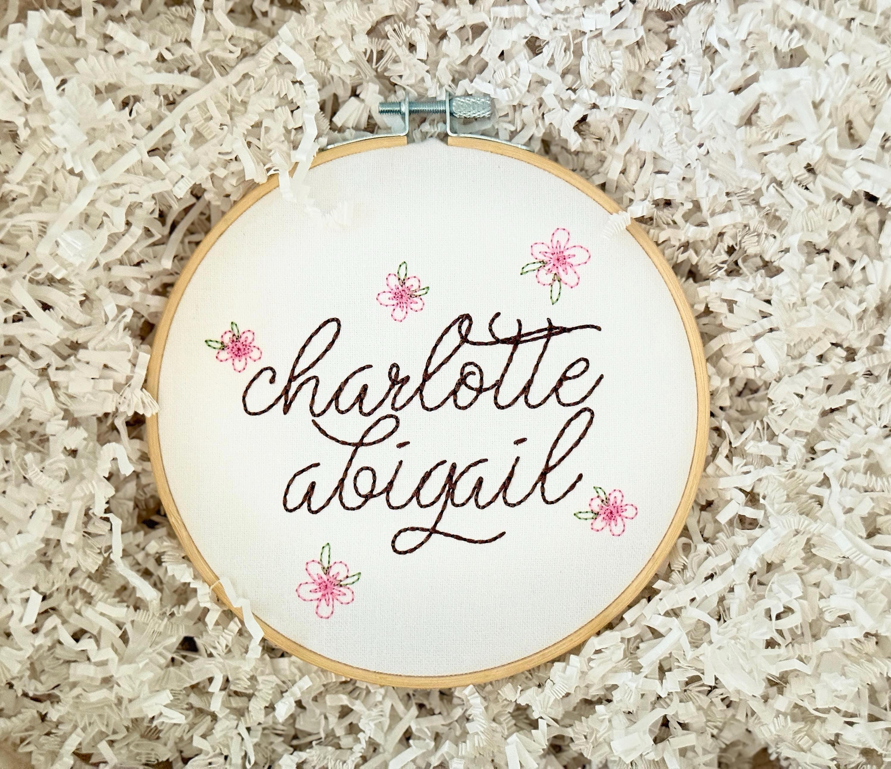 Light wood embroidery hoop art set on a bed of white crinkle paper with the name Charlotte Abigail embroidered in dark brown. The hoop has five pink embroidered flowers around the name: Charlotte Abigail in cursive. It is stitched on white cotton fabric.