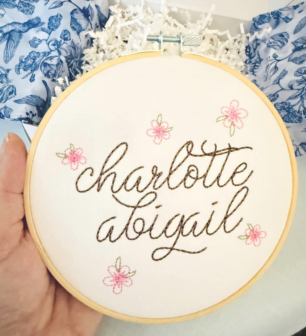 A light wood embroidery hoop art being held in one hand. The hoop has five pink embroidered flowers around the name: Charlotte Abigail in cursive. It is stitched on white cotton fabric.