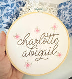 A light wood embroidery hoop art being held in one hand. The hoop has five pink embroidered flowers around the name: Charlotte Abigail in cursive. It is stitched on white cotton fabric.